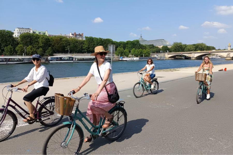 Four women cycling along the Seine riverbank on a sunny day, with the Grand Palais visible in the background, Paris