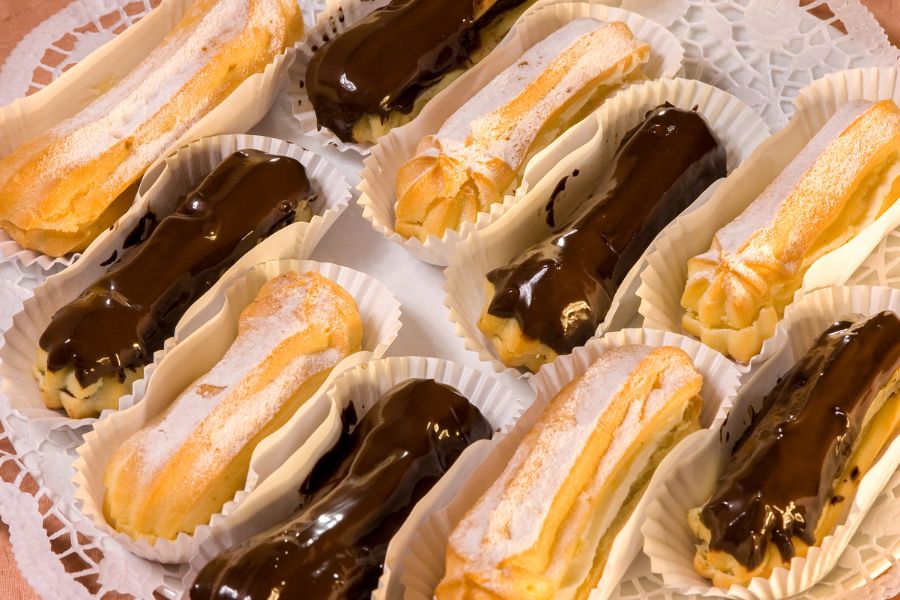 A tray of French éclairs in paper cases, some topped with glossy dark chocolate ganache and others dusted with icing sugar and filled with cream