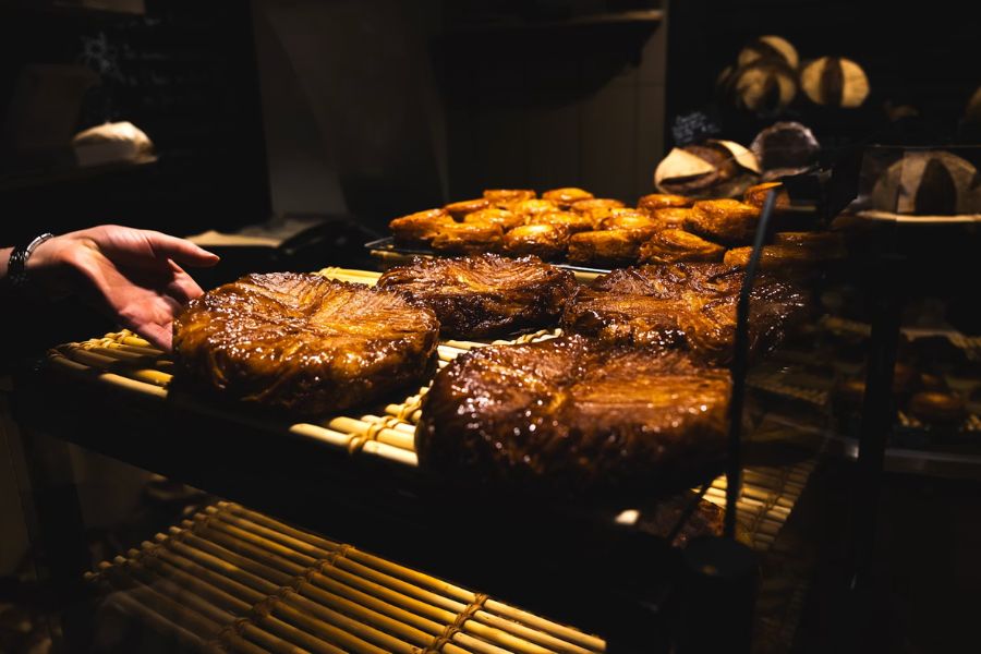 A bakery rack holding large, deeply caramelised kouign-amann pastries with a glossy, lacquered surface, with a hand reaching in to select one