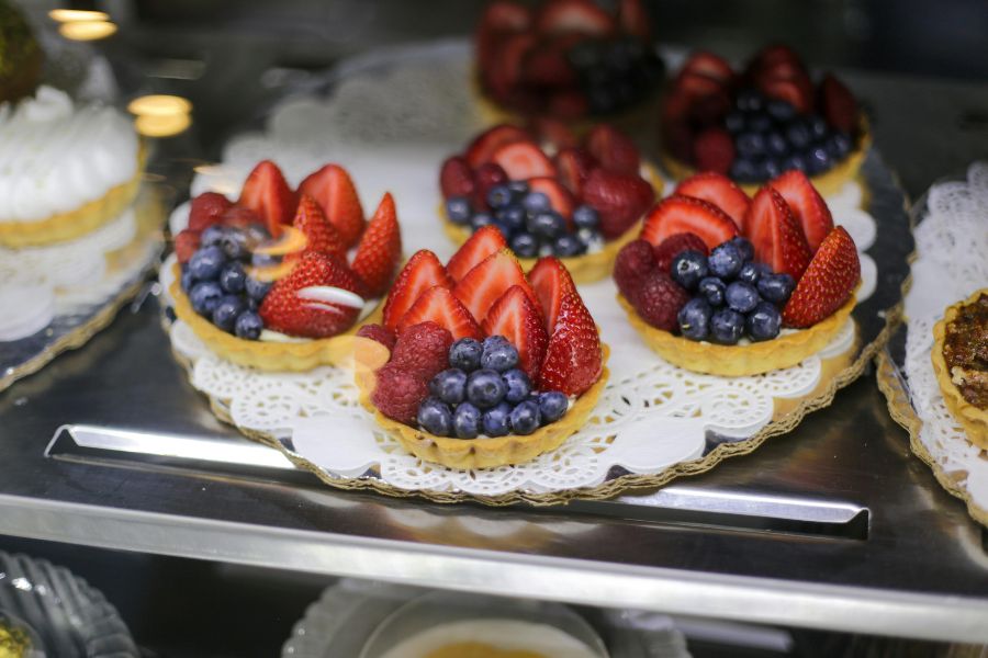 Individual fruit tarts displayed in a patisserie case, each filled with crème pâtissière and topped with fresh strawberries, blueberries and raspberries on a pastry base