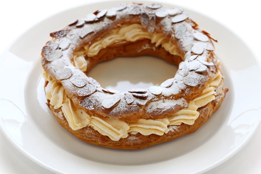A Paris-Brest on a white plate — a ring of choux pastry filled with praline cream and topped with flaked almonds and a dusting of icing sugar