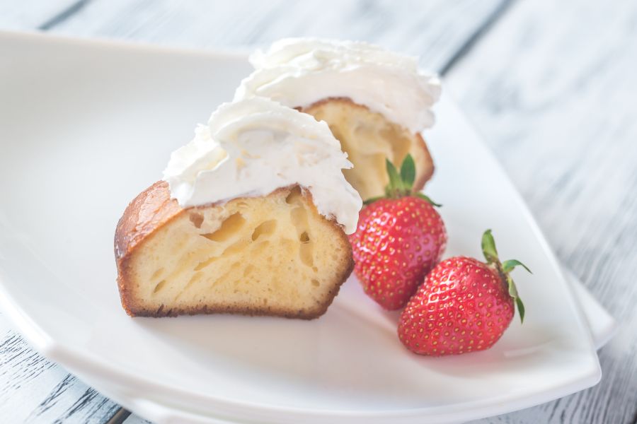 A halved savarin or baba au rhum on a white plate, showing its airy, spongy texture, topped with whipped cream and served with fresh strawberries
