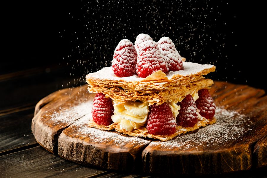 A mille-feuille with layers of flaky puff pastry and vanilla cream, topped with fresh raspberries and a dusting of icing sugar falling from above, photographed against a dark background