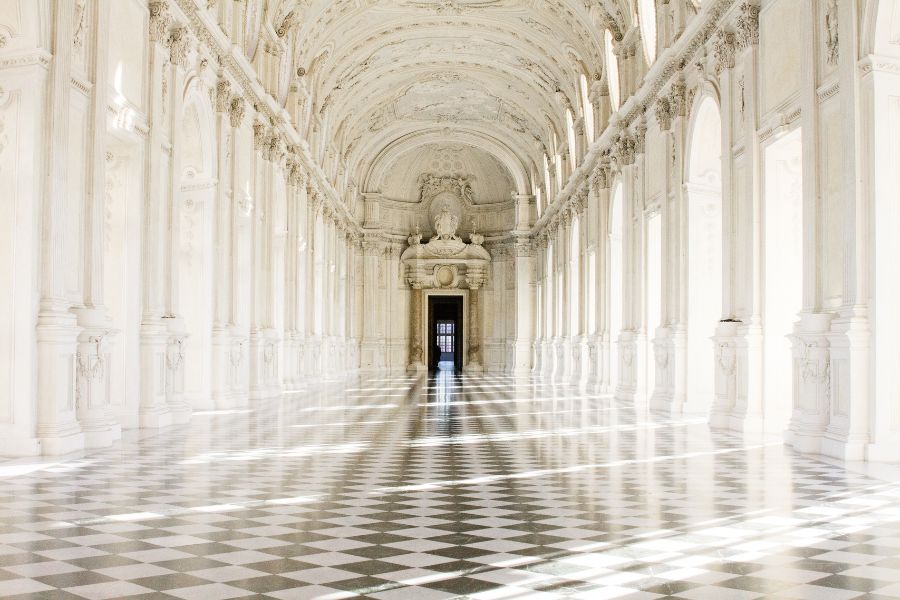 This is an image of a long, fancy hallway in a palace with checkered marble tiles and pillars leading to another room.
