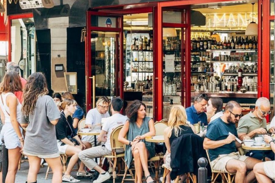 This is an image of a very busy cafe with people sitting inside and on seats along the sidewalk.