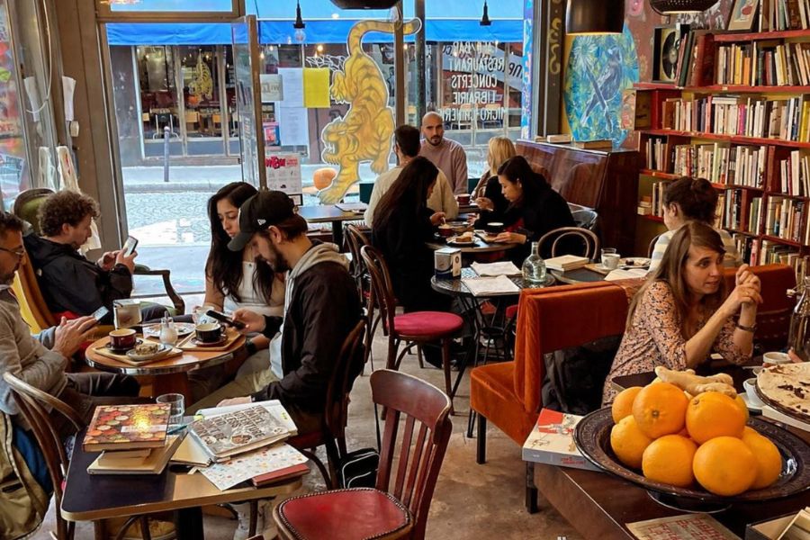 Best Non-Touristy Cafes in Paris: This is an image of the cosy interior of a little café with plenty of people chatting with friends and reading books. There is also a book shelf in the corner.