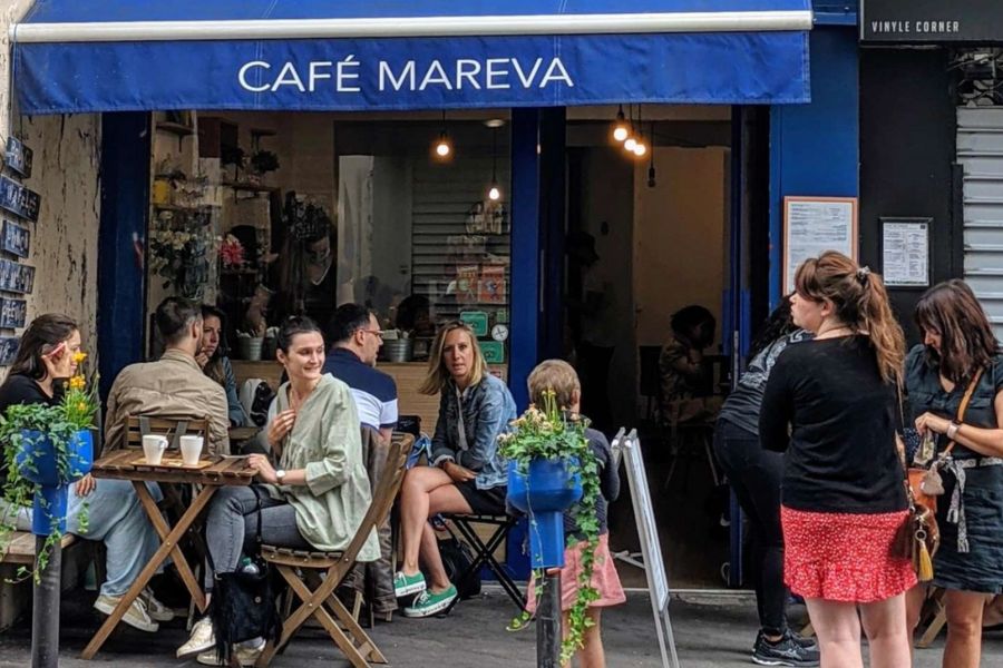 This is an image of the exterior of a Parisian cafe with dark blue accents and lots of people sitting on tables outside eating and drinking.