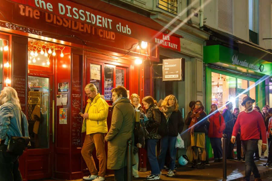 This is a picture of a jazz bar from the exterior road side. People are seen lining up to get inside.