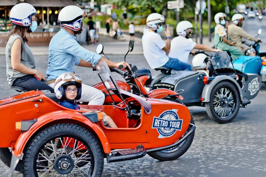 A convoy of vintage red, black, and blue sidecar motorcycles on a Paris street, riders and passengers wearing retro helmets