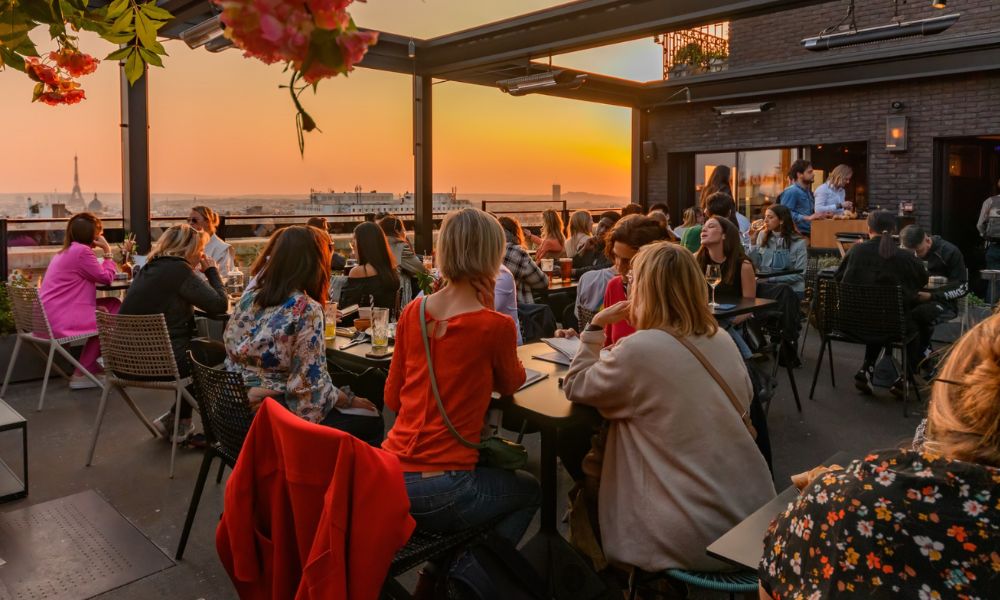 This is an image of a rooftop full of people sitting on different tables with a view of Paris's skyline and the sunset.