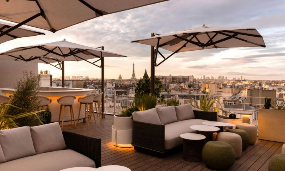 This is an image of a rather swanky rooftop bar with cream furniture and the city in the distance with shades, couches and tables.
