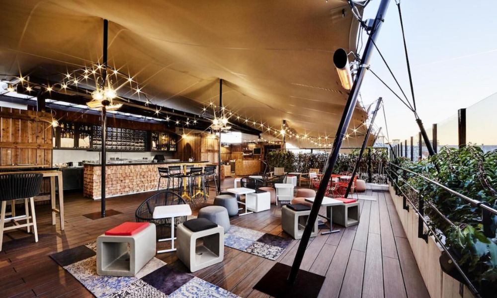 This is an image of a nice rooftop bat with cushioned seating, a boho-looking bar and fun fairy lights draped around.