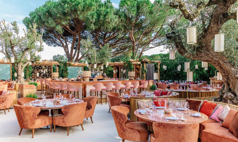 This is an image of an outdoor balcony with a bunch of greenery and lots of orange tables.