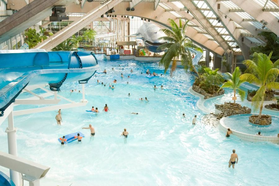 An indoor water park with a large wave pool, water slides, and palm trees beneath a vaulted glass roof