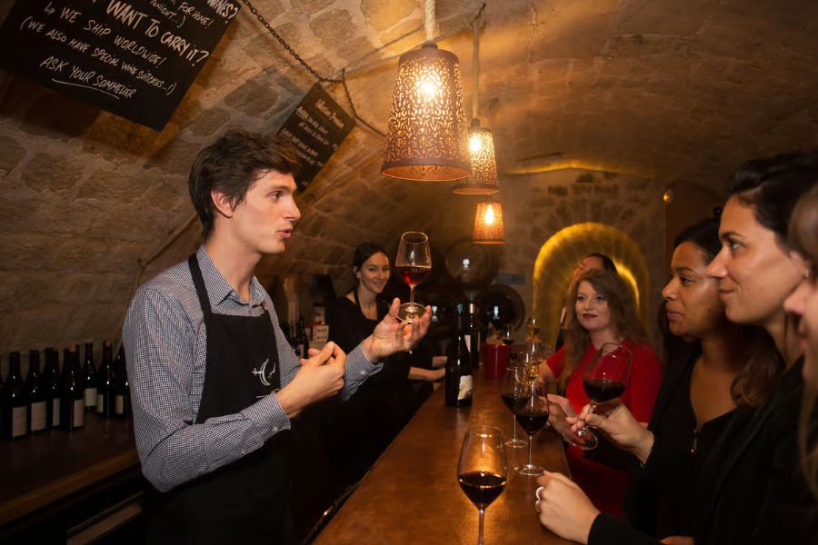 A sommelier leading a wine tasting in a candlelit stone cellar, holding a glass of red wine up to a small group of guests