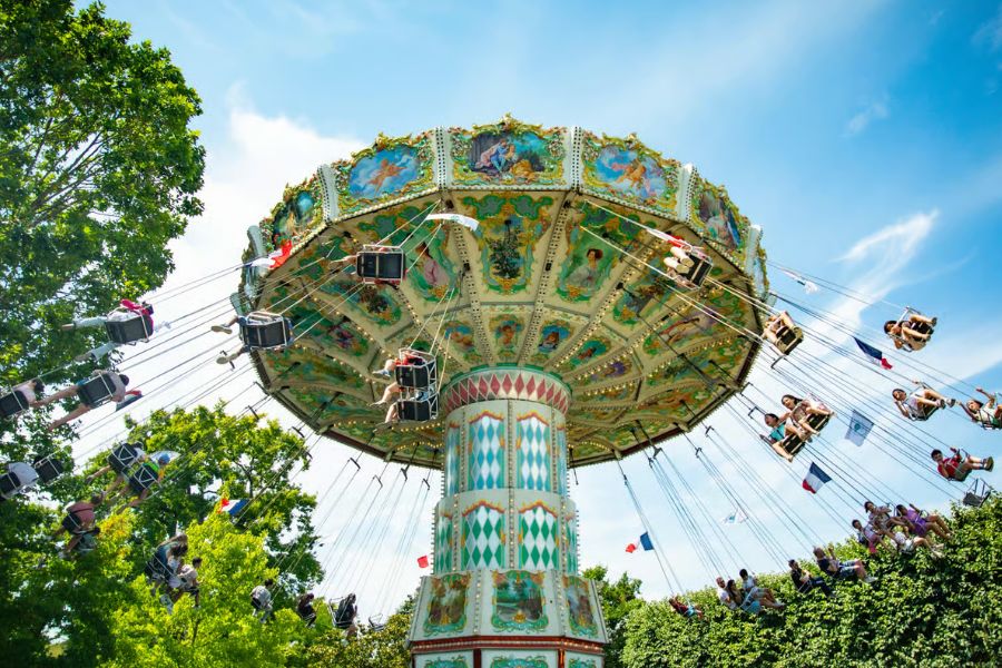 A ornate vintage carousel with swing seats, decorated with painted cherubs and French flags, photographed from below against a blue sky