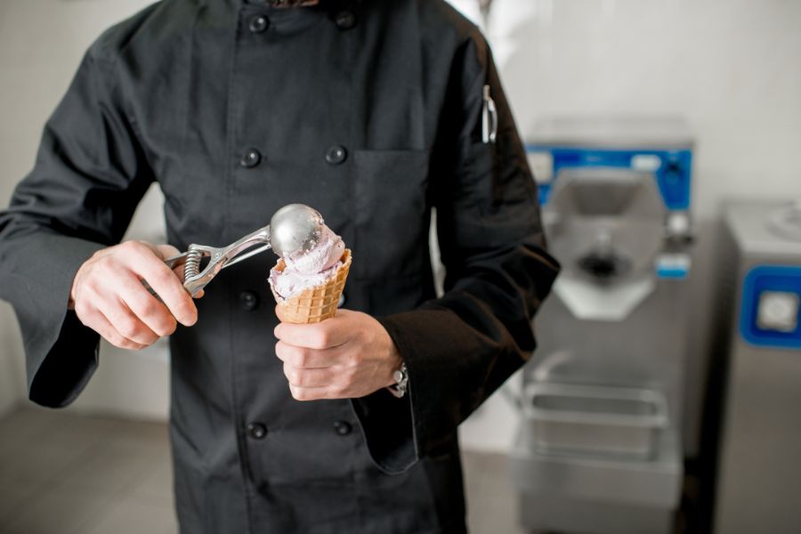 A chef in black whites scooping a ball of pale purple ice cream into a waffle cone