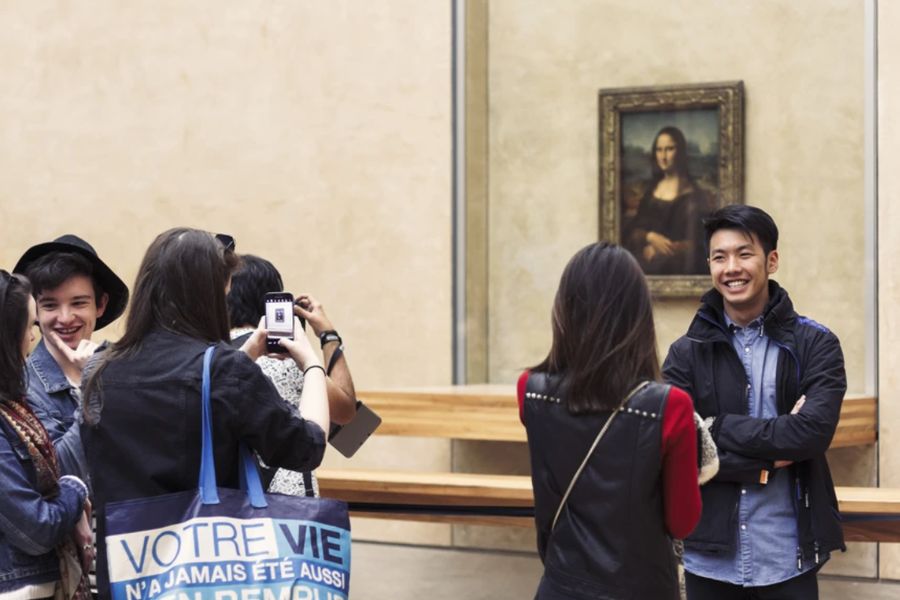 Tourists photographing the Mona Lisa at the Louvre museum, with the famous painting visible behind a barrier in the background