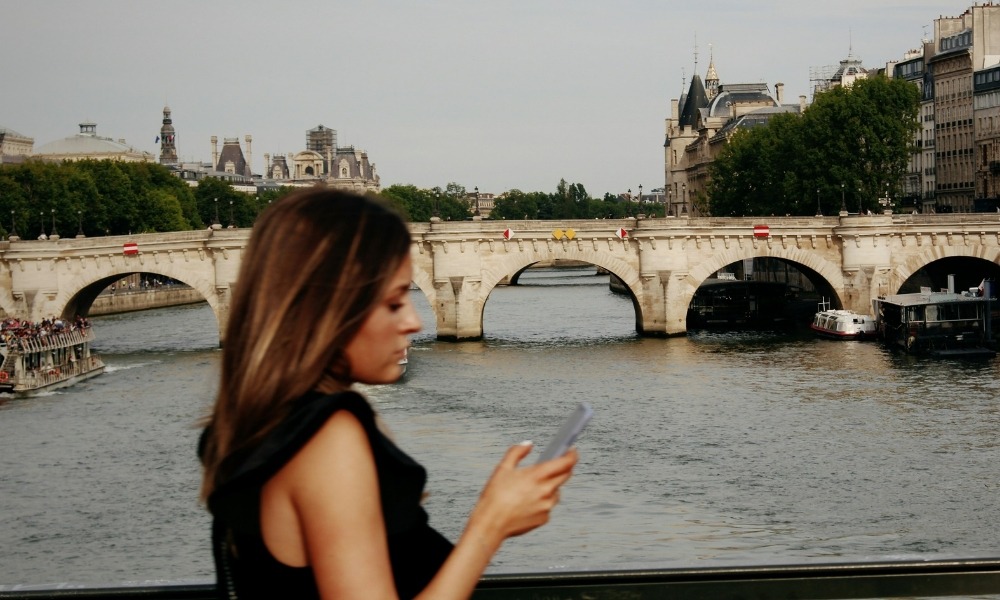 A woman holding a phone walks along a bridge in Paris.