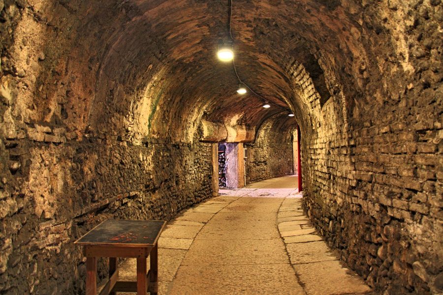 A dimly lit stone tunnel with arched ceiling and flagstone floor, lit by overhead bulbs receding into the distance