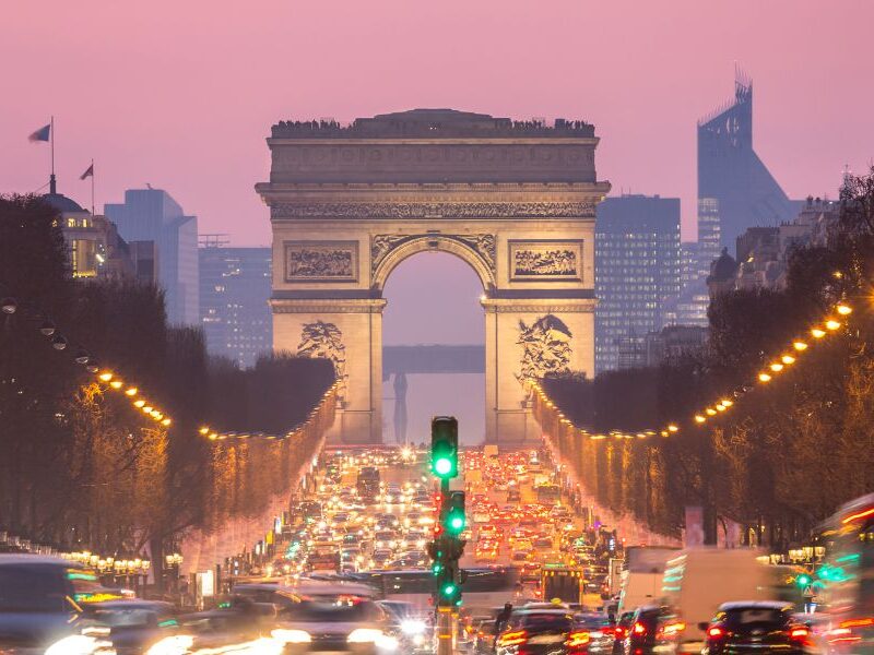 This is an image of the Champs Elysees with the Arc De Triomphe in the distance. The streets are full of cars and there is a pink sunset.
