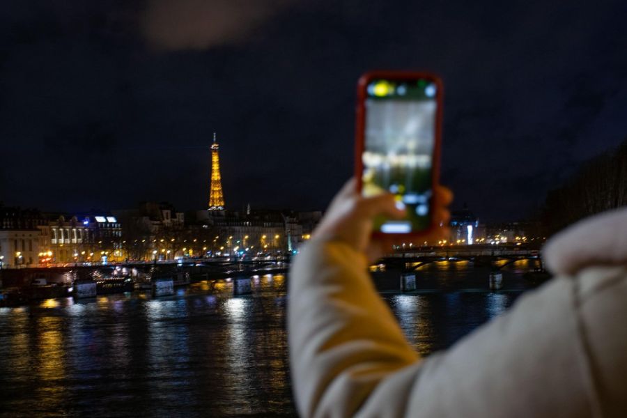 A person holding up a smartphone to photograph the illuminated Eiffel Tower at night, reflected in the Seine river below