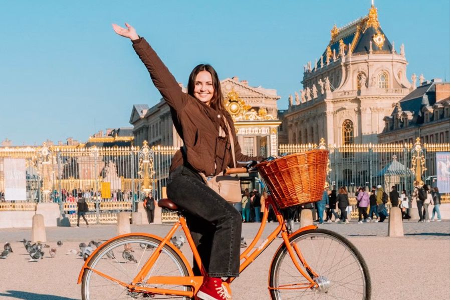A smiling woman on an orange bicycle with a wicker basket, arm raised, in front of the gilded gates of the Palace of Versailles