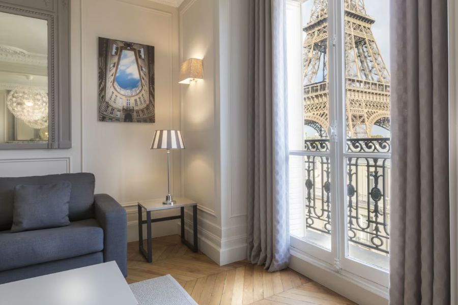 This is a minimalist apart-hotel in Paris of a living room couch with a side table lamp and art. The window also looks out on to the Eiffel Tower.