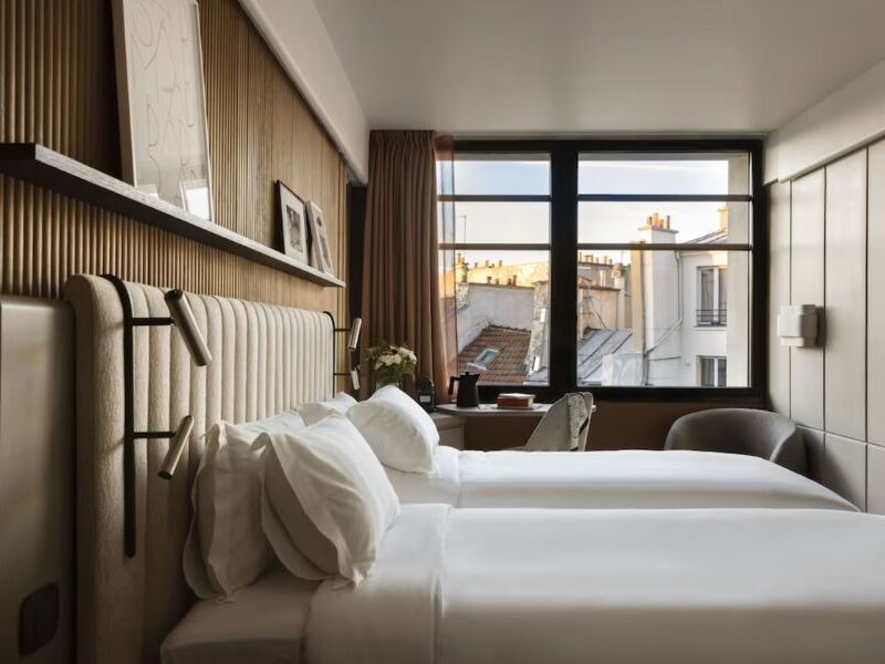 This is an image of a minimalist hotel room with beige features and two single beds.