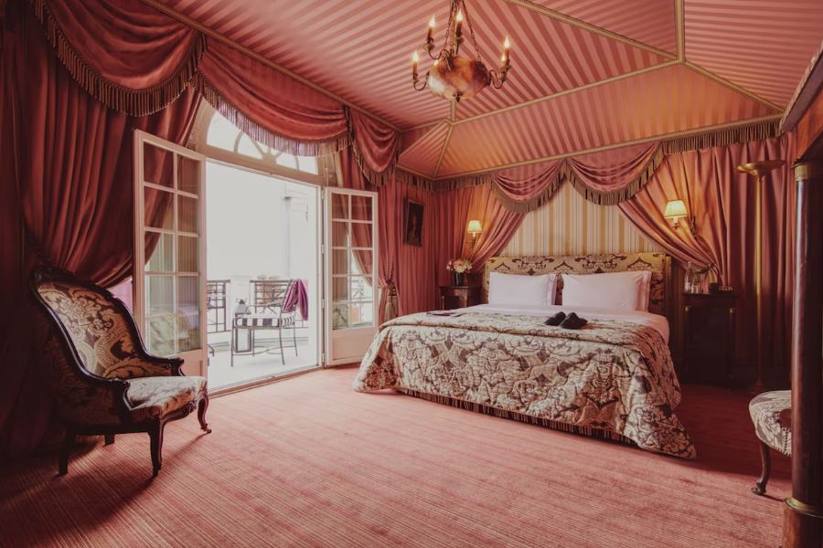 This is a very regal hotel room with red walls and moody lamp lighting and a double bed.