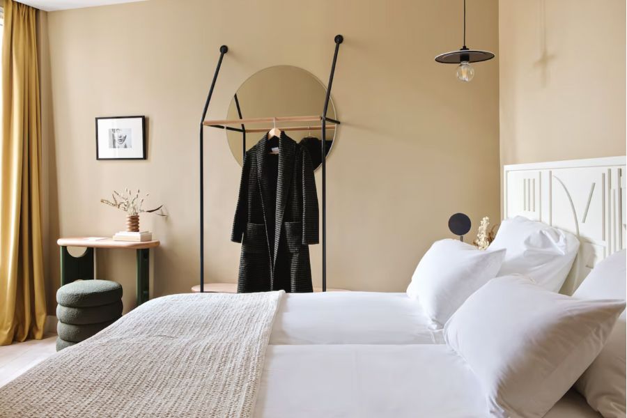 This is a cool, minimalist hotel room in the Latin Quarter of Paris featuring white bedding and beige walls. A black robe hangs in the middle of the room.