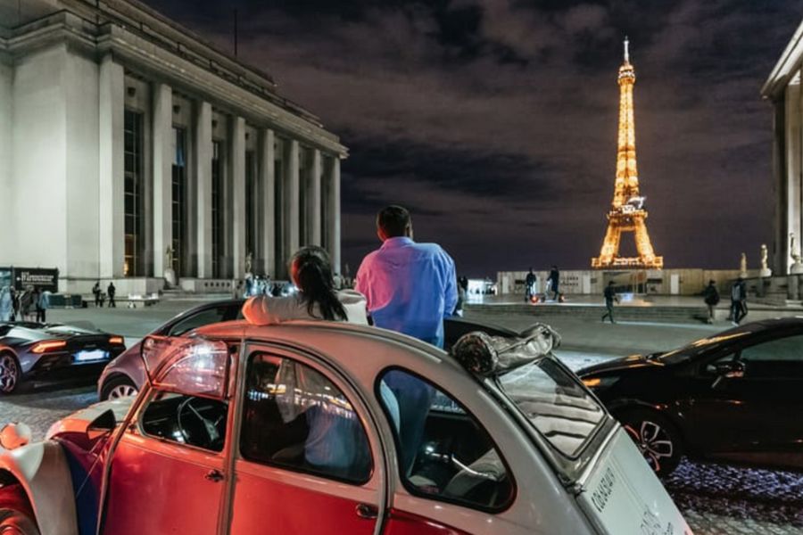 This is an image of a couple on a tour around Paris in a vintage car. They are parked along Trocadéro and can see the Eiffel Tower in the distance.