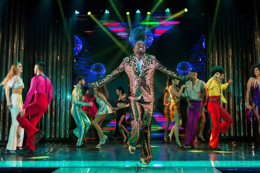 This is an image of a bunch of performers on stage in bright colours and sparkly suits dancing around theatrically.