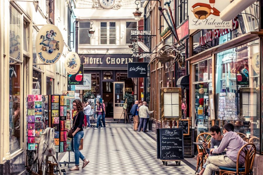 This is a picture of a small shopping passage in Paris with small shops and people walking and looking out front.