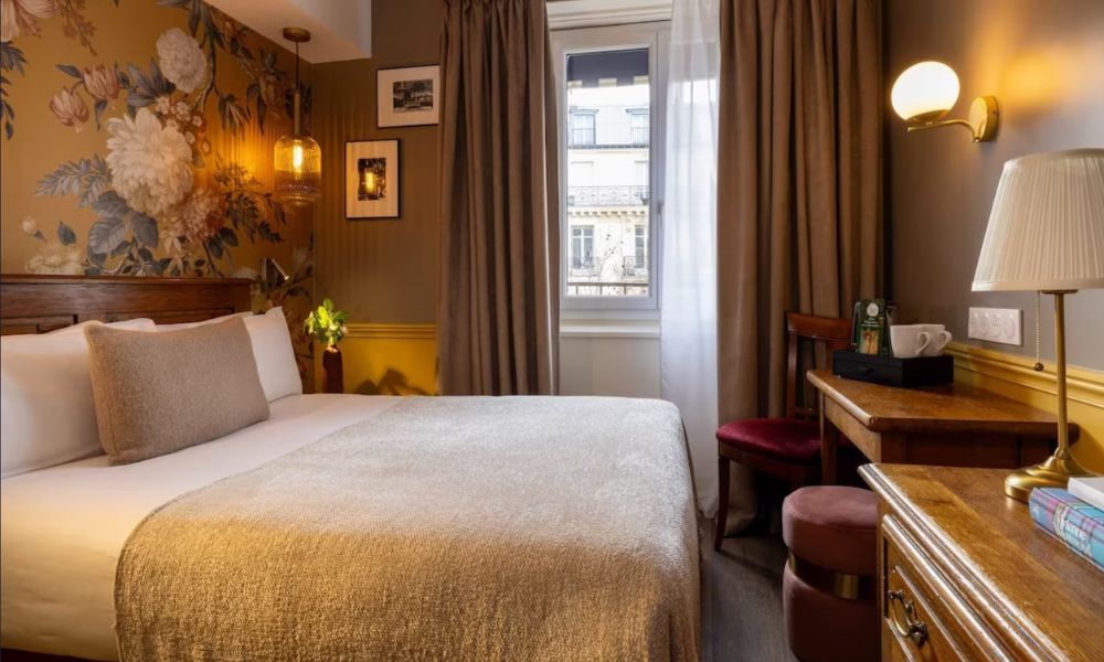 This is an image of a small hotel room with a small window and double bed. There is also a desk and small vanity. It is dark, but the lamps and mood lighting make up for it.