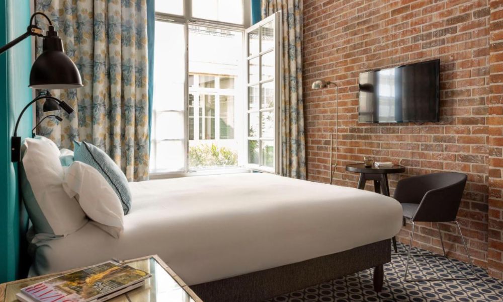Best Paris hotels for solo travelers: This is an image of a nice hotel room with exposed brick interiors, a double bed and a large window that lets in so much light.