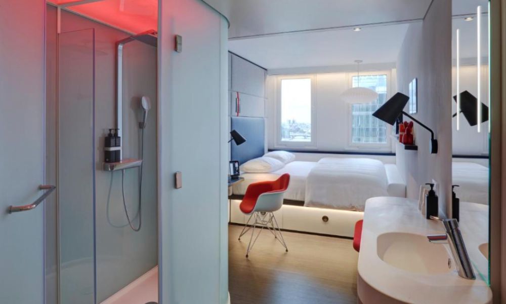 This is an image of a large, minimalist hotel room with a desk, double bed, and small open shower.