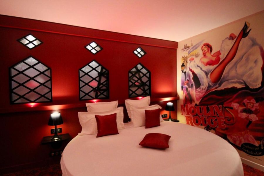 This is an image of a red hotel room with a bed that is circular in shape. There is a Moulin Rouge artwork on the wall.