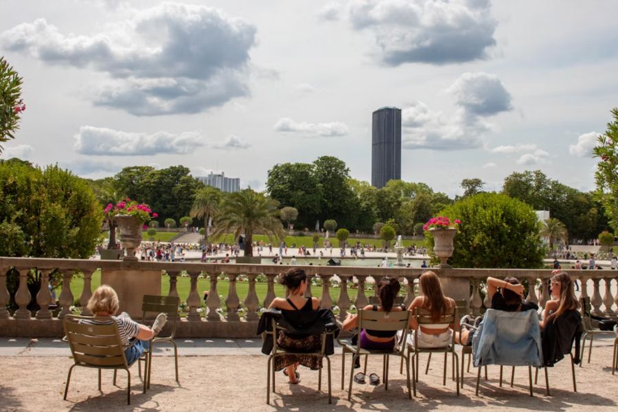 People relaxing in green metal chairs on a terrace overlooking the grand central fountain and manicured lawns of Jardin du Luxembourg, with Tour Montparnasse visible in the background on a sunny summer's day in Paris.