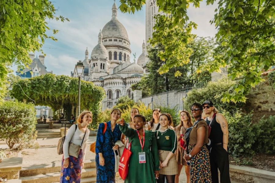 A tour guide pointing out landmarks to a smiling group of visitors on a walking tour in Montmartre, with the white stone domes of Sacré-Cœur Basilica rising through the trees behind them.