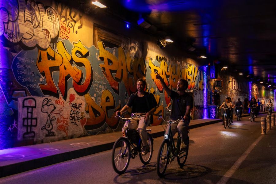 Two cyclists riding through a graffiti-covered underpass at night, surrounded by bold orange and yellow street art, as other riders follow in the background on a Paris night bike tour.