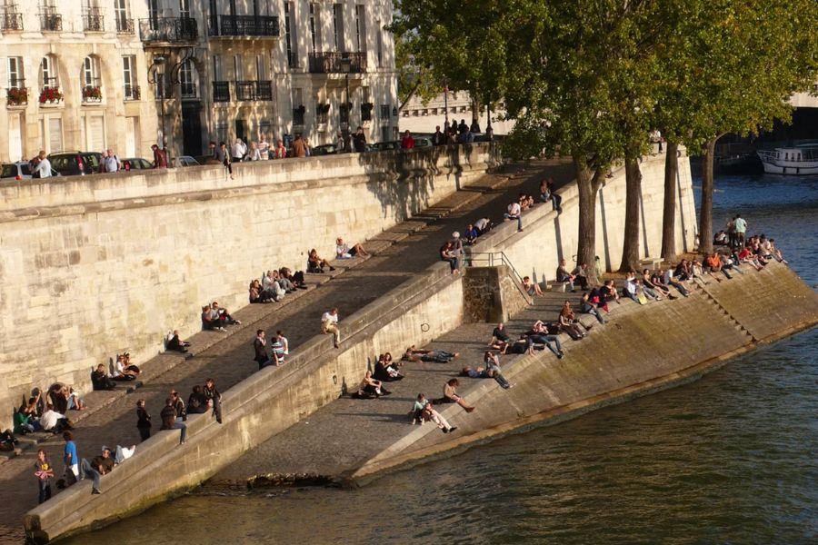 Crowds of Parisians sitting and lounging on the stone steps and quaysides along the Seine riverbank in golden afternoon light.