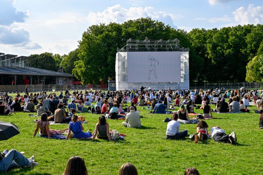 A large crowd sitting on grass at an outdoor cinema screening in a Paris park on a bright summer afternoon, with a giant screen showing an animated film.