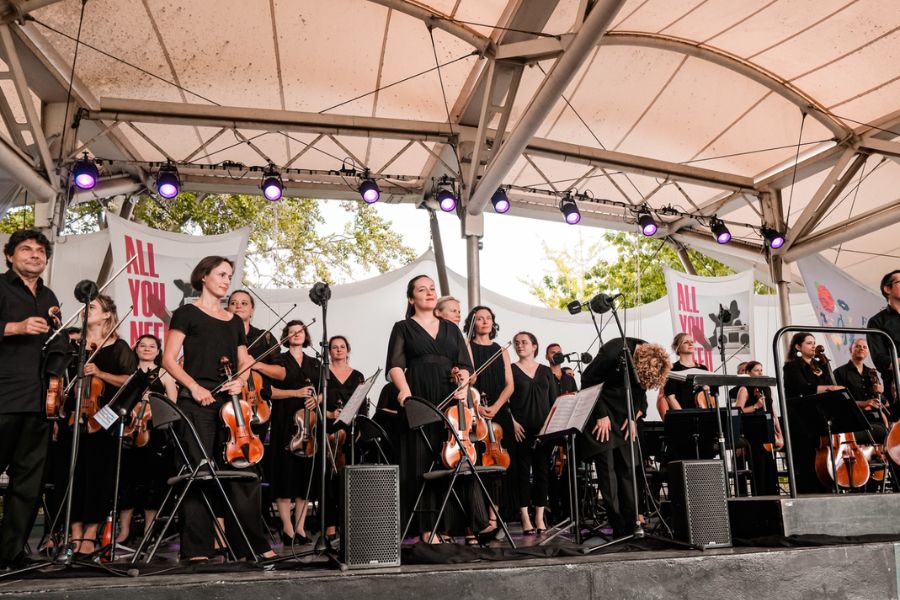An orchestra performing an outdoor concert in Paris in August, with musicians dressed in black holding string instruments on a covered open-air stage.