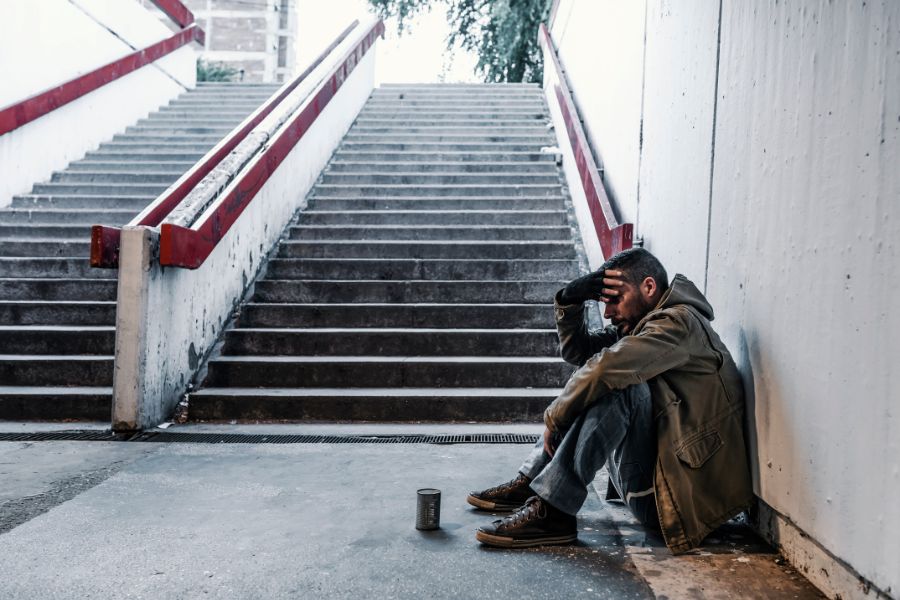 This is an image of a homeless man sitting by the bottom of the stairs on a sidewalk with a cup in front of him.