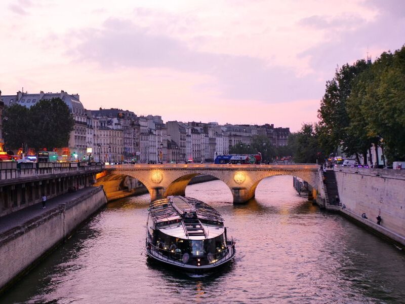 This is an image of the Seine River at sunset with a boat cruising through and a bridge in the background.