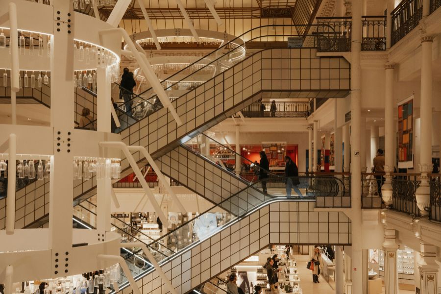 Interior of Le Bon Marché department store in Paris, showing multiple floors connected by criss-crossing escalators, ornate white ironwork columns, warm lighting, and shoppers browsing beauty and fragrance counters on the ground floor.