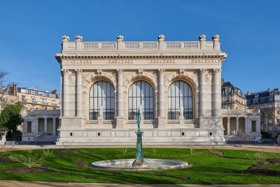 The neoclassical facade of the Palais Galliera fashion museum in Paris on a clear blue sky day, featuring three large arched windows, ornate stone columns, sculpted details, and a bronze fountain surrounded by green lawn in the foreground.