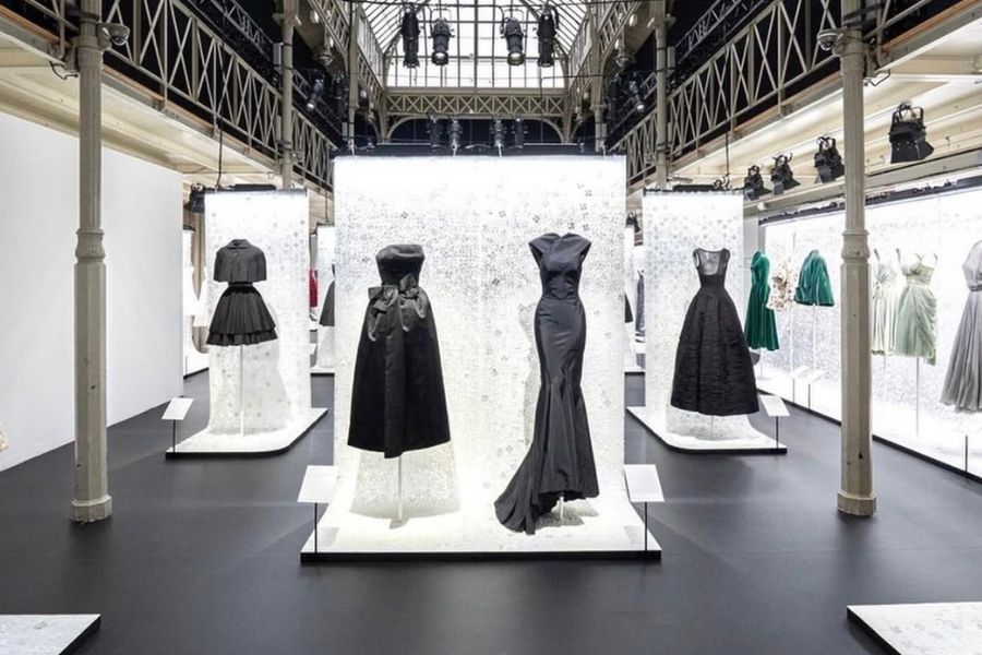 A fashion exhibition space with black flooring and an industrial glass ceiling, displaying multiple haute couture gowns on illuminated white pedestals, including dramatic black evening gowns and colourful dresses on either side.