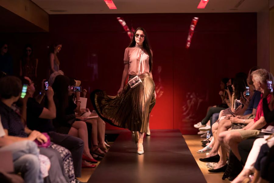 A model walking a fashion show runway wearing a sheer blush top, flowing metallic gold pleated midi skirt, sunglasses, and white boots, with an audience photographing on smartphones on both sides against a deep red backdrop.